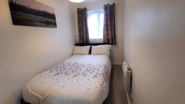 A small, single-bed room with a painting on the wall, a window with curtains, and a radiator. - Joydens Wood, 2-bed Apt, Sleeps 4 With Parking (Grays)