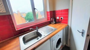 A compact kitchen with a window, a sink, a washing machine, and a red-tiled backsplash. - Joydens Wood, 2-bed Apt, Sleeps 4 With Parking (Grays)