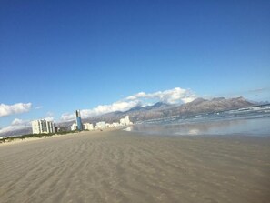 A beach with buildings in the background and mountains in the distance. - A gorgeous studio with mountain views at Goedehoop, Strand, Western Cape (Cape Town)
