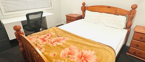 Une chambre à coucher avec un lit en bois, un bureau et une chaise. Les murs sont ornés d’œuvres d’art et il y a une fenêtre avec des stores.