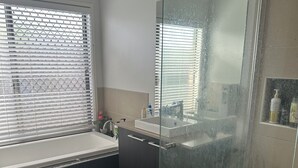 A modern bathroom with a glass shower enclosure, a window with blinds, a sink, and a small shelf with toiletries.