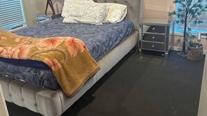 Une chambre à coucher avec une tête de lit capitonnée, un lit avec une literie à motifs, une table de chevet avec une lampe et une plante décorative.