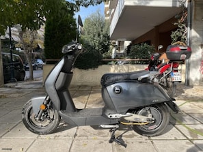 Ein Roller steht auf befestigter Fläche, im Hintergrund sind ein Gebäude und Bäume zu sehen.