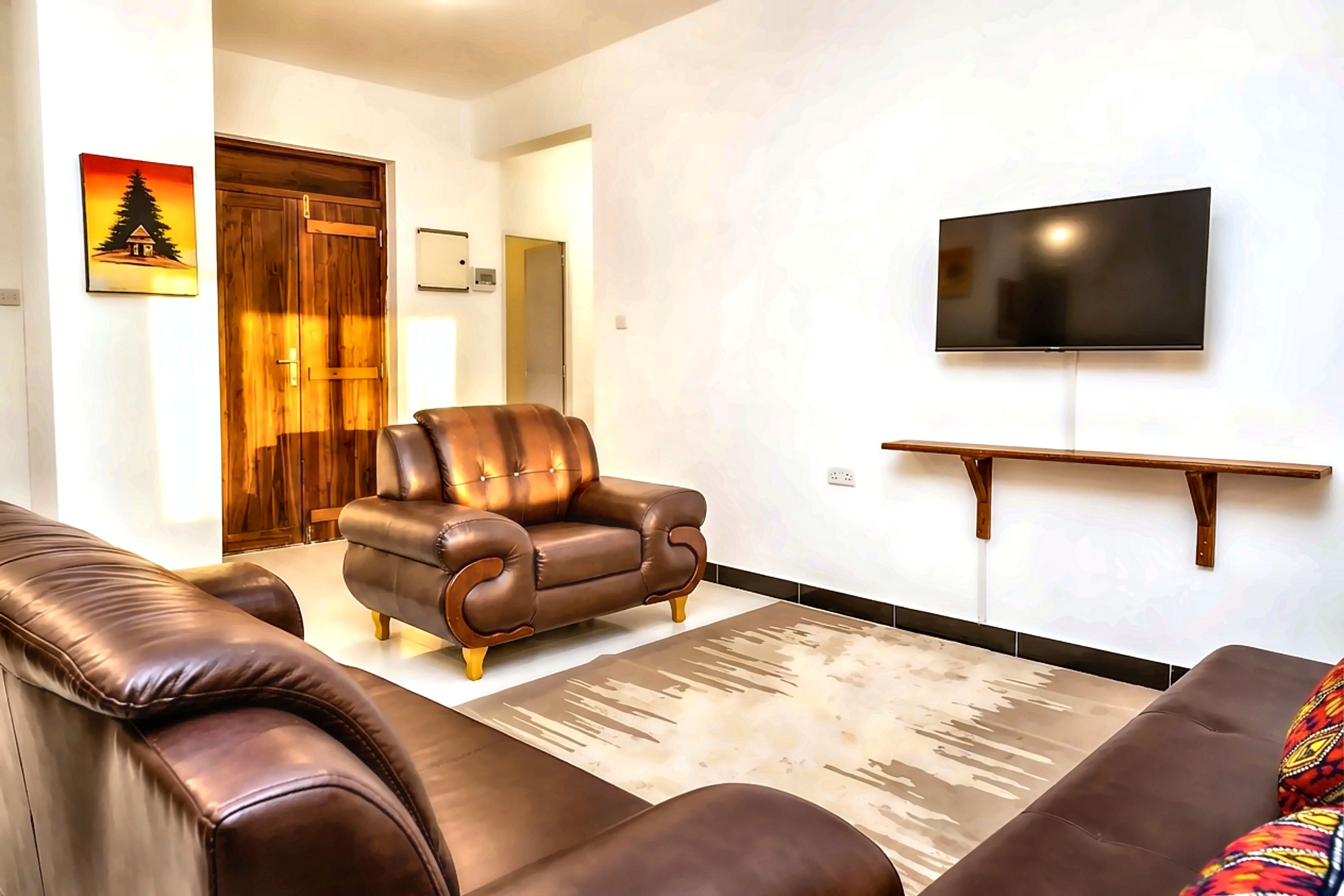 A living room with a brown leather sofa, a matching armchair, a flat-screen TV mounted on the wall, and a wooden door with a picture of a mountain.