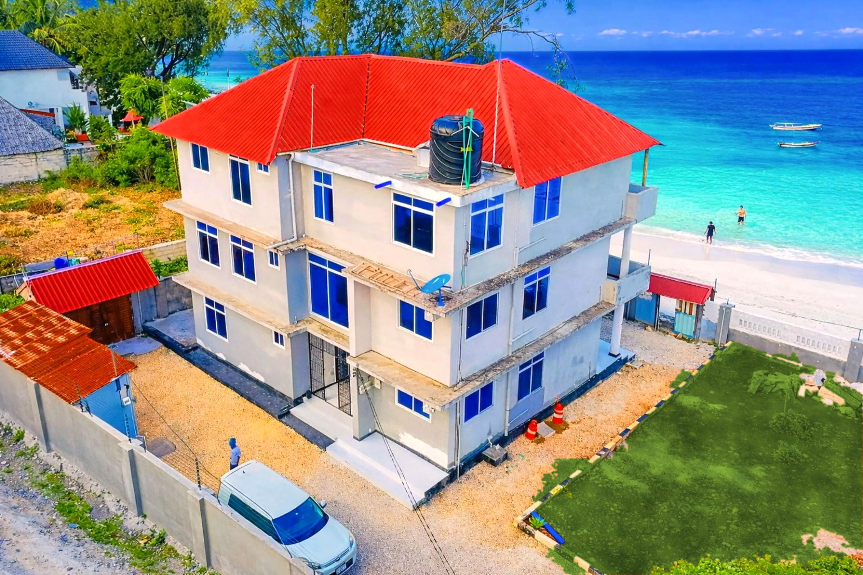 A multi-story building with a red roof, located near a beach with clear blue water.