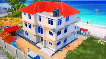 A multi-story building with a red roof, located near a beach with clear blue water.