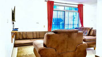 A living room with a brown leather sofa, a brown armchair, a TV mounted on the wall, and a window with red curtains.