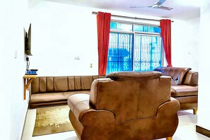A living room with a brown leather sofa, a brown armchair, a TV mounted on the wall, and a window with red curtains. - Exclusive Beachfront Apartment in Uroa, Zanzibar - Ocean Views (Zanzibar, Uroa)