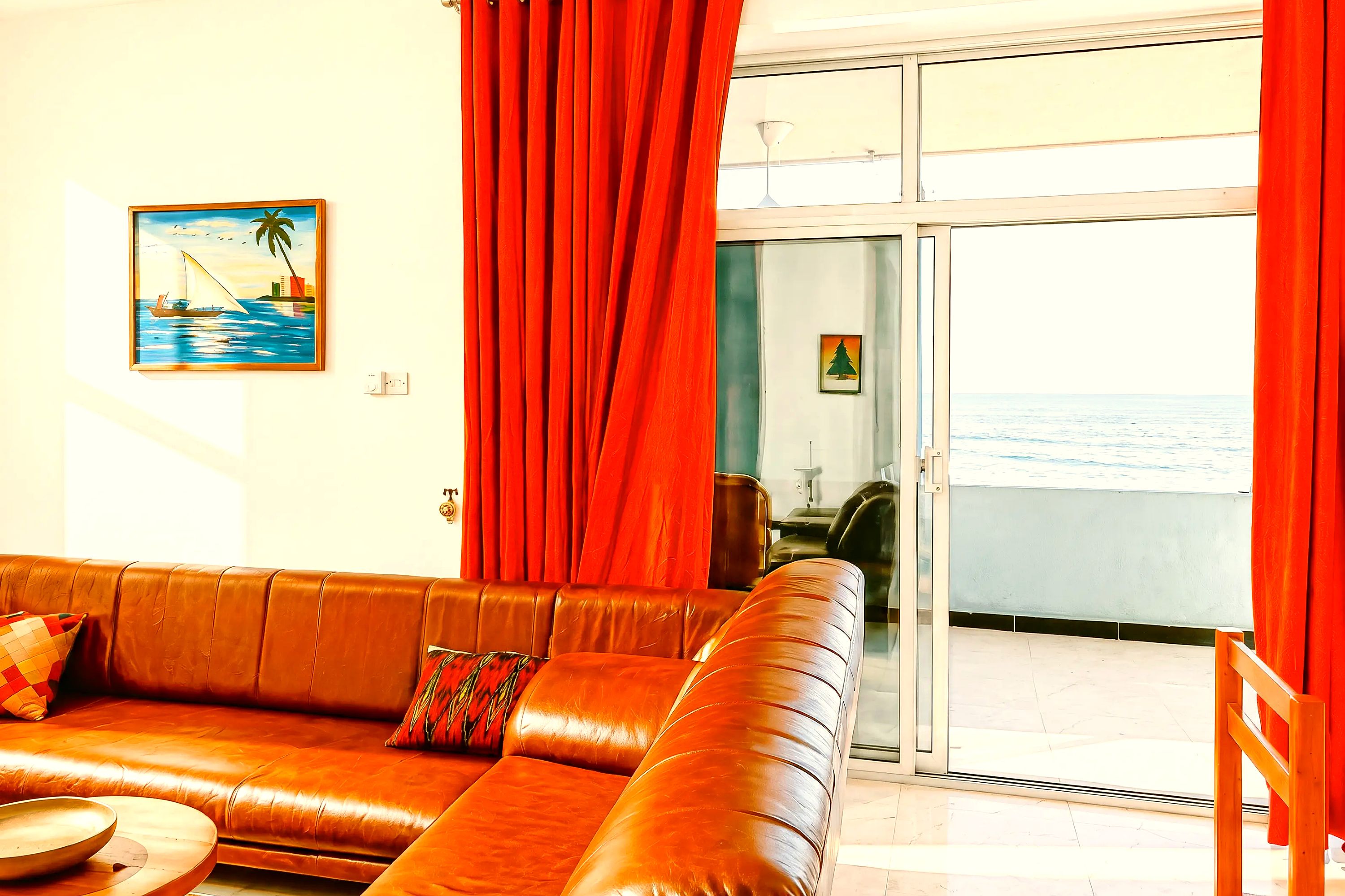 A living room with a brown leather sofa, a painting of a sailboat, and a view of the sea through a glass door.
