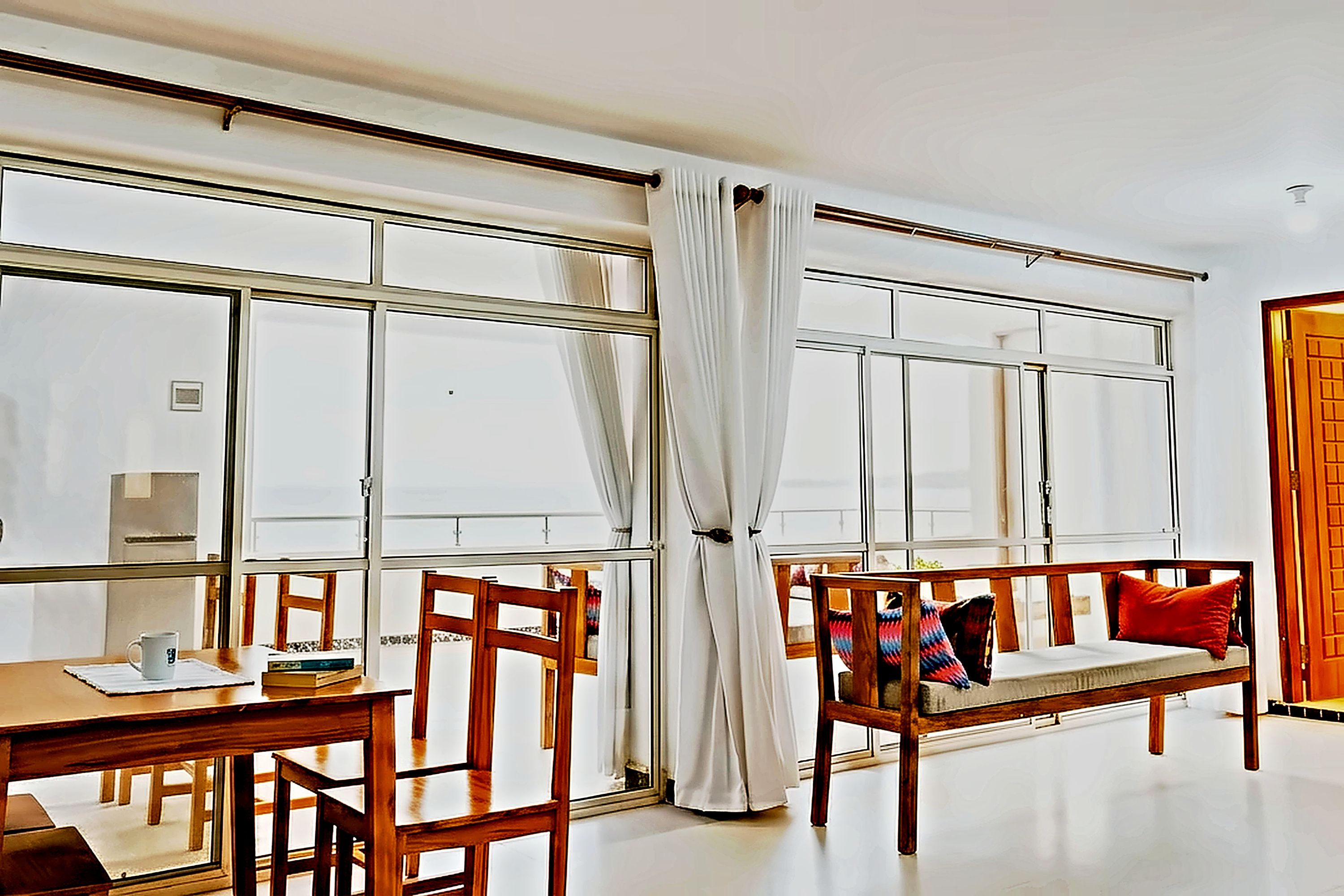 Una habitación con un ventanal grande, un banco de madera con cojines, una mesa con una taza y libros, y una puerta con marco de madera.