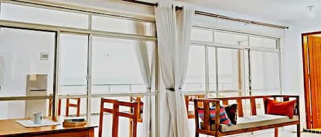 Una habitación con un ventanal grande, un banco de madera con cojines, una mesa con una taza y libros, y una puerta con marco de madera.