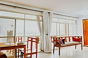 Ein Zimmer mit einem großen Fenster, einer hölzernen Bank mit Kissen, einem Tisch mit Tasse und Büchern sowie einer Tür mit Holzrahmen.