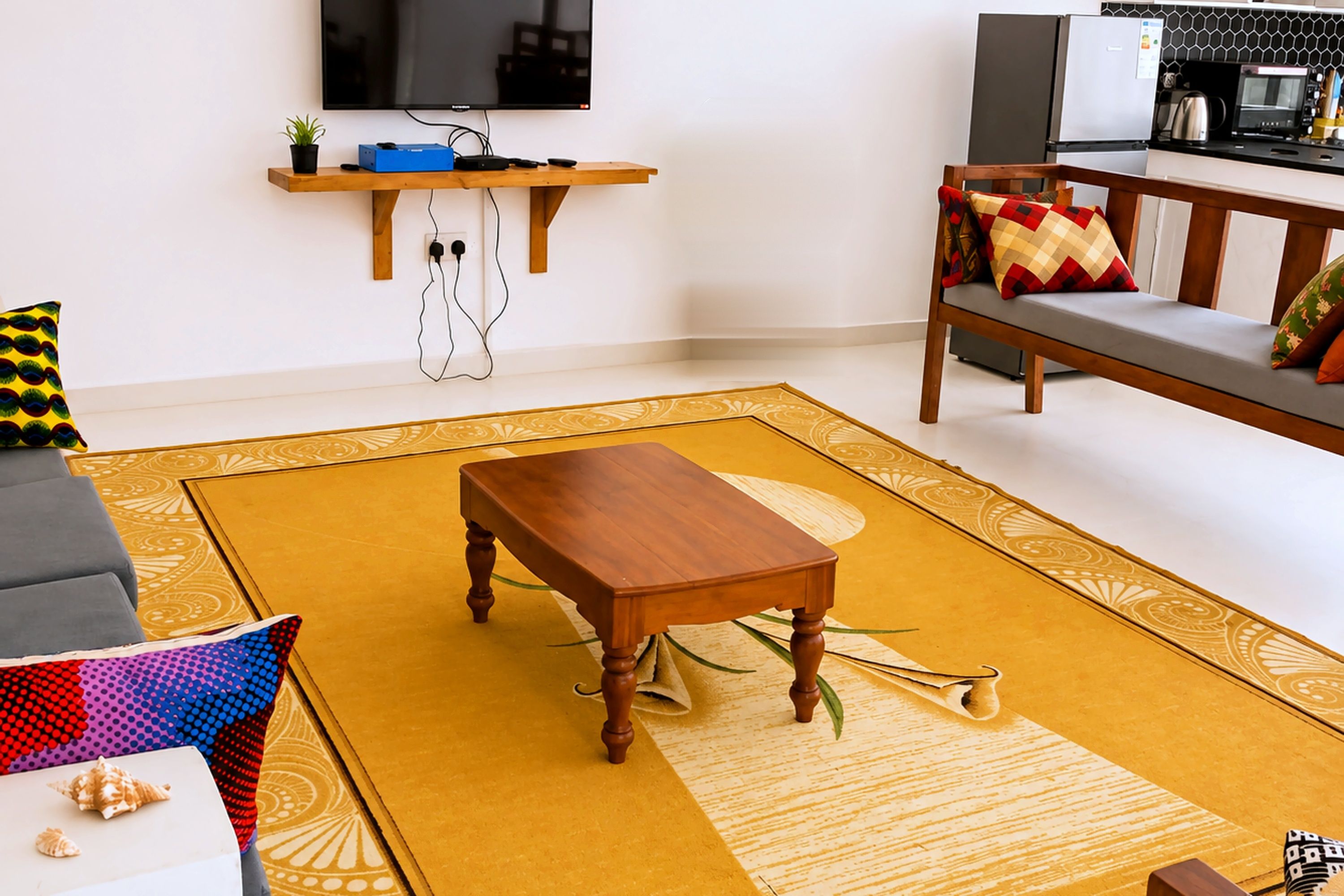 Una sala de estar con una mesa de centro de madera, un sofá con cojines estampados, un televisor de pantalla plana montado en la pared y una zona de cocina al fondo.
