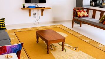 Una sala de estar con una mesa de centro de madera, un sofá con cojines estampados, un televisor de pantalla plana montado en la pared y una zona de cocina al fondo.
