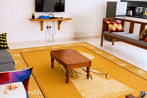 Un salon avec une table basse en bois, un canapé agrémenté de coussins à motifs, un téléviseur à écran plat fixé au mur et, au fond, un coin cuisine.