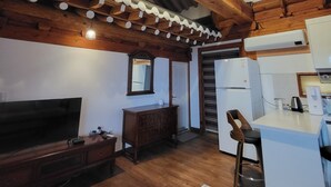 A rustic interior with exposed wooden beams, a television, a wooden dresser, and a kitchen area with a refrigerator. - Hanok Pension Daega (Jeju City)