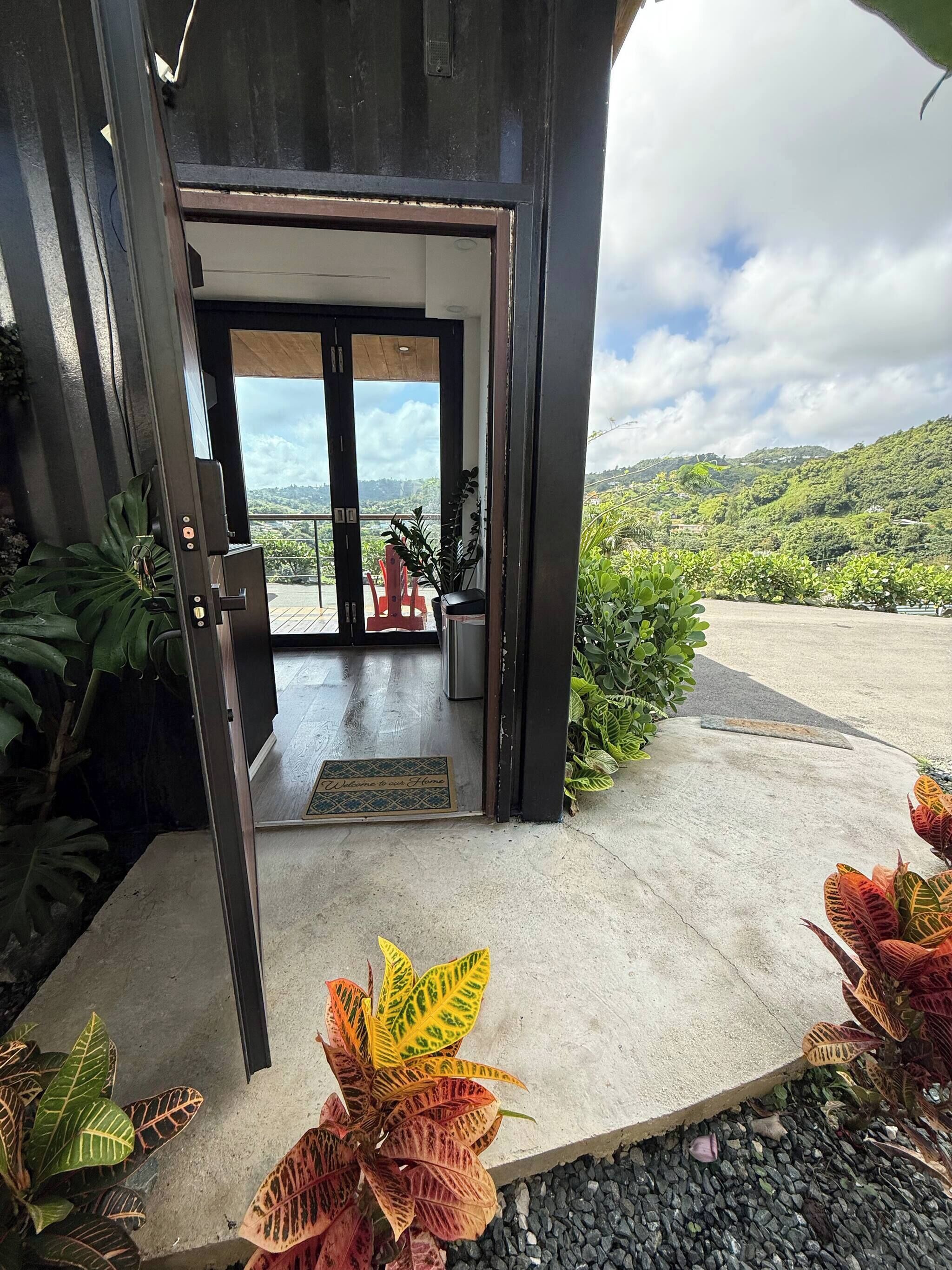 Une entrée moderne avec une porte en verre, un paillasson et une vue sur un paysage verdoyant.