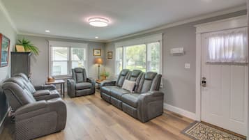 A living room with a sectional sofa, a chair, a small table, and a lamp. There are windows with blinds, a door with a curtain, and a ceiling light.