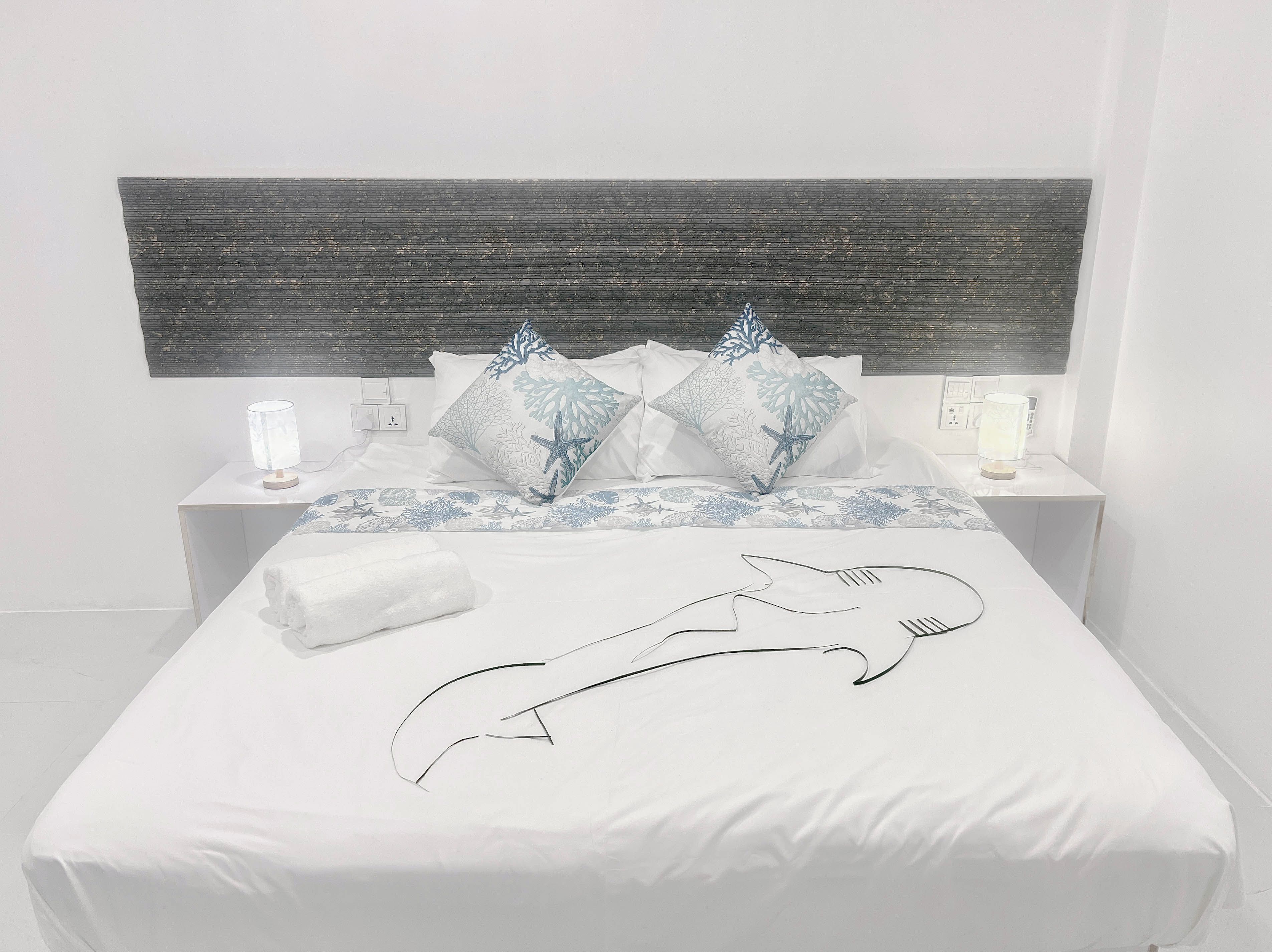 Una cama con cubre cama blanco, dos mesitas de noche con lámparas y un cabecero con diseño texturizado.