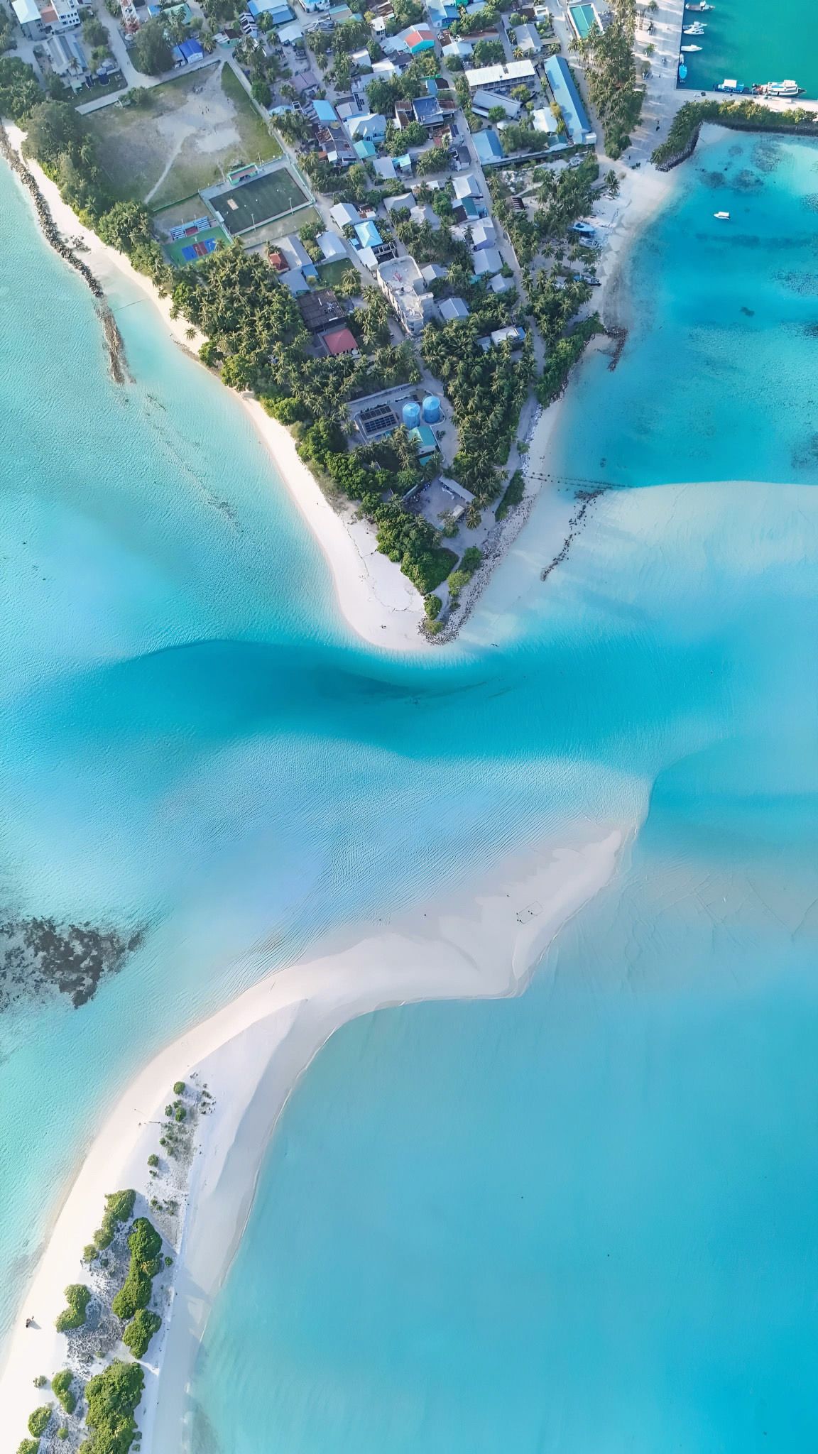 An aerial view of a coastal area with a beach, clear turquoise waters, and a residential area with buildings.