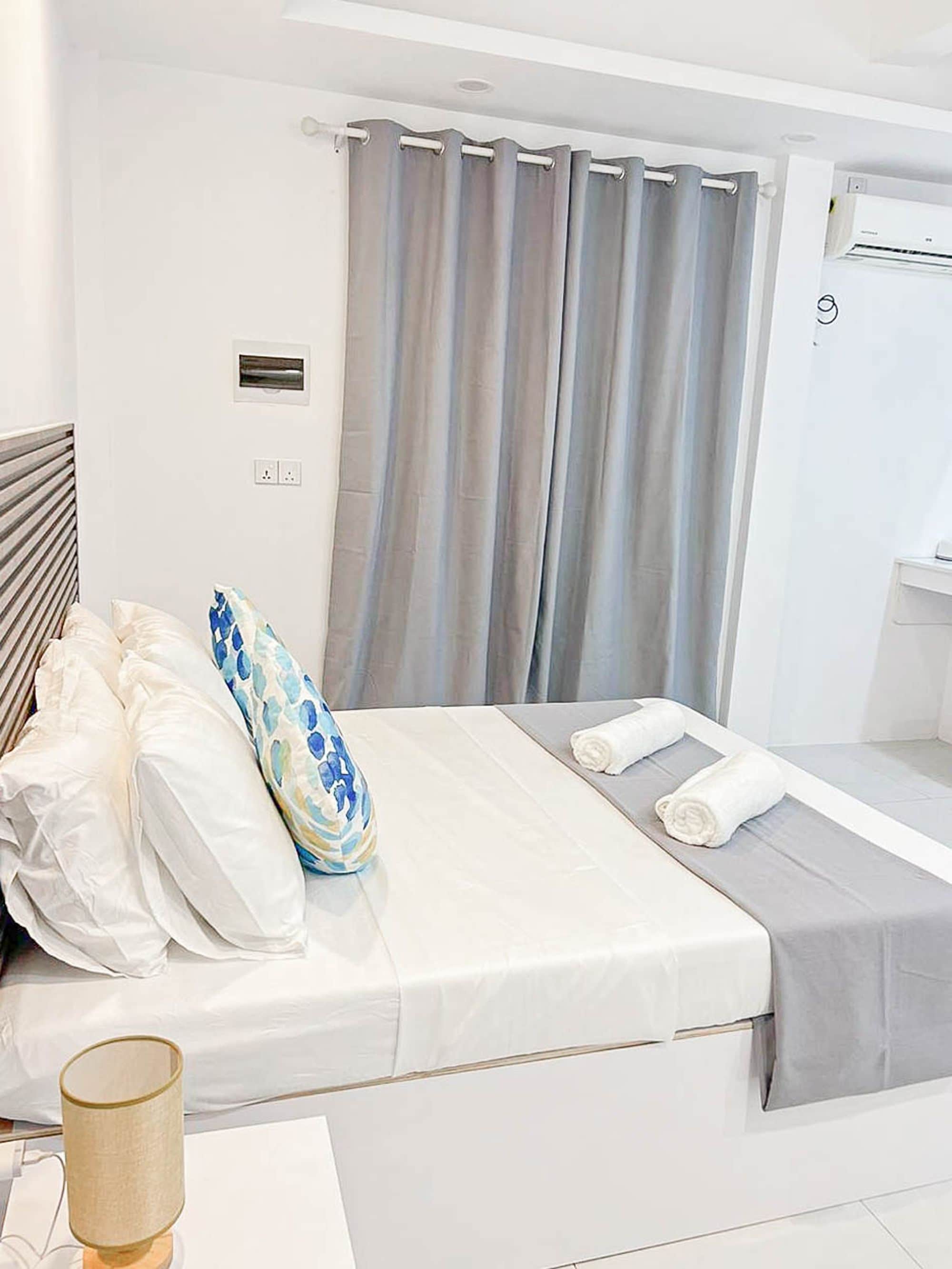 Habitación de hotel con cama, mesita de noche, lámpara, ventana con cortinas y aire acondicionado.