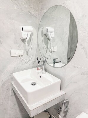 A bathroom with a white sink, a round mirror, two hair dryers mounted on the wall, and a toilet.