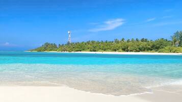 A beach with clear turquoise water, white sand, and a small island with dense green vegetation and a tall structure.