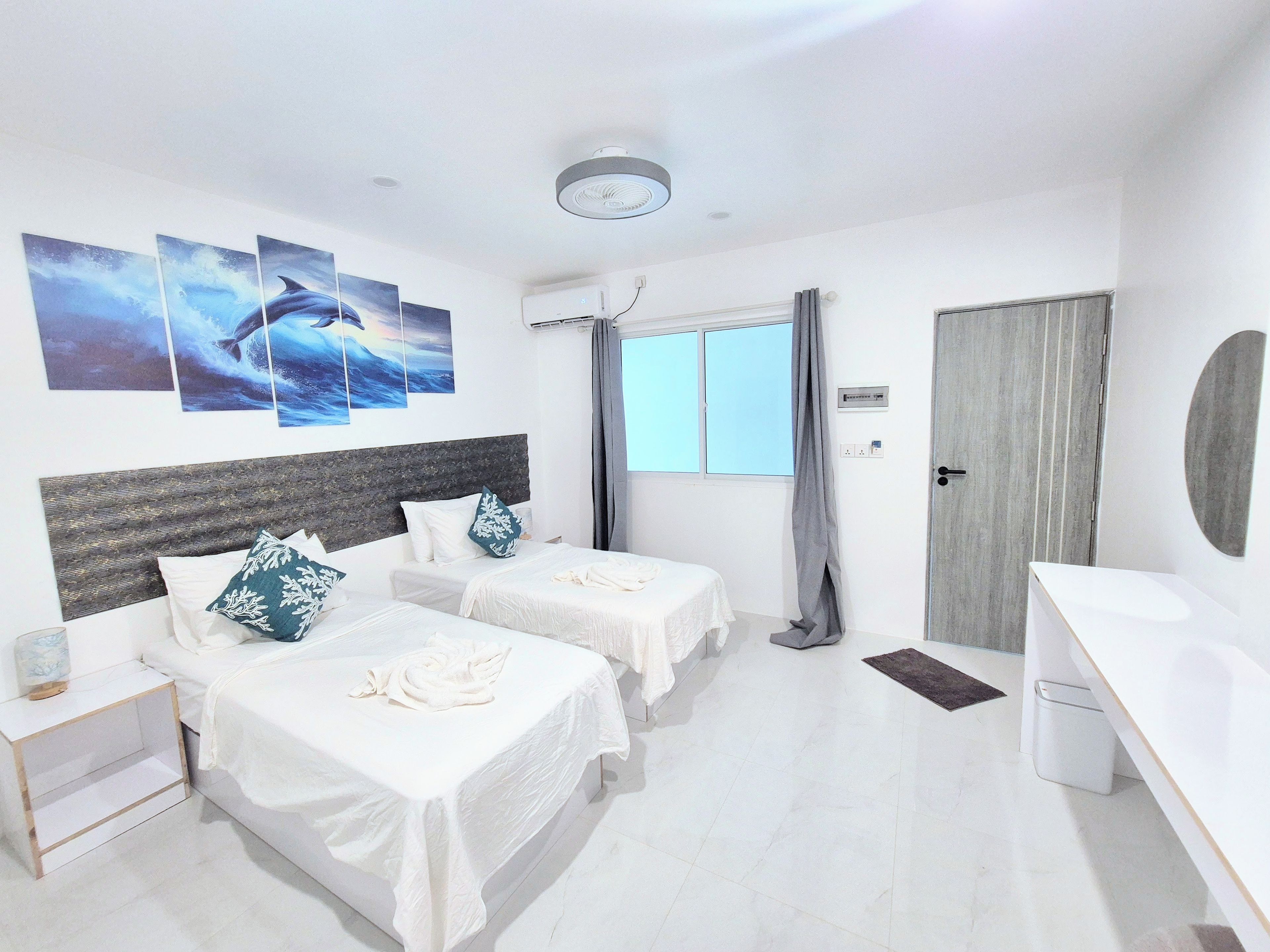 A hotel room with two beds, a desk, a window with curtains, and artwork on the wall.