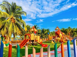 Un parque infantil con toboganes y estructuras para escalar, rodeado de palmeras y un cielo despejado.