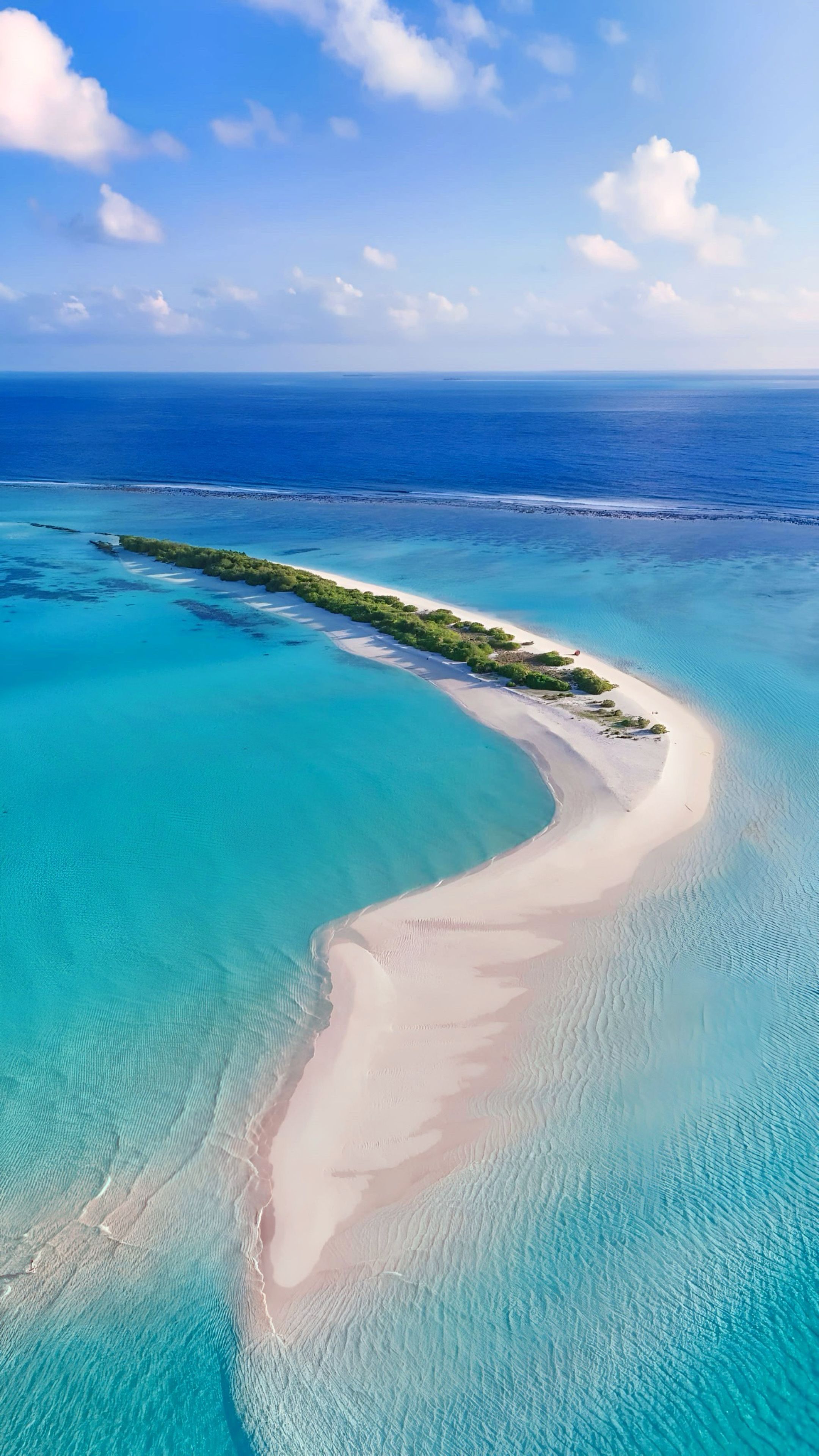 An aerial view of a coastal area with clear turquoise waters, a sandy beach, and a narrow strip of vegetation.