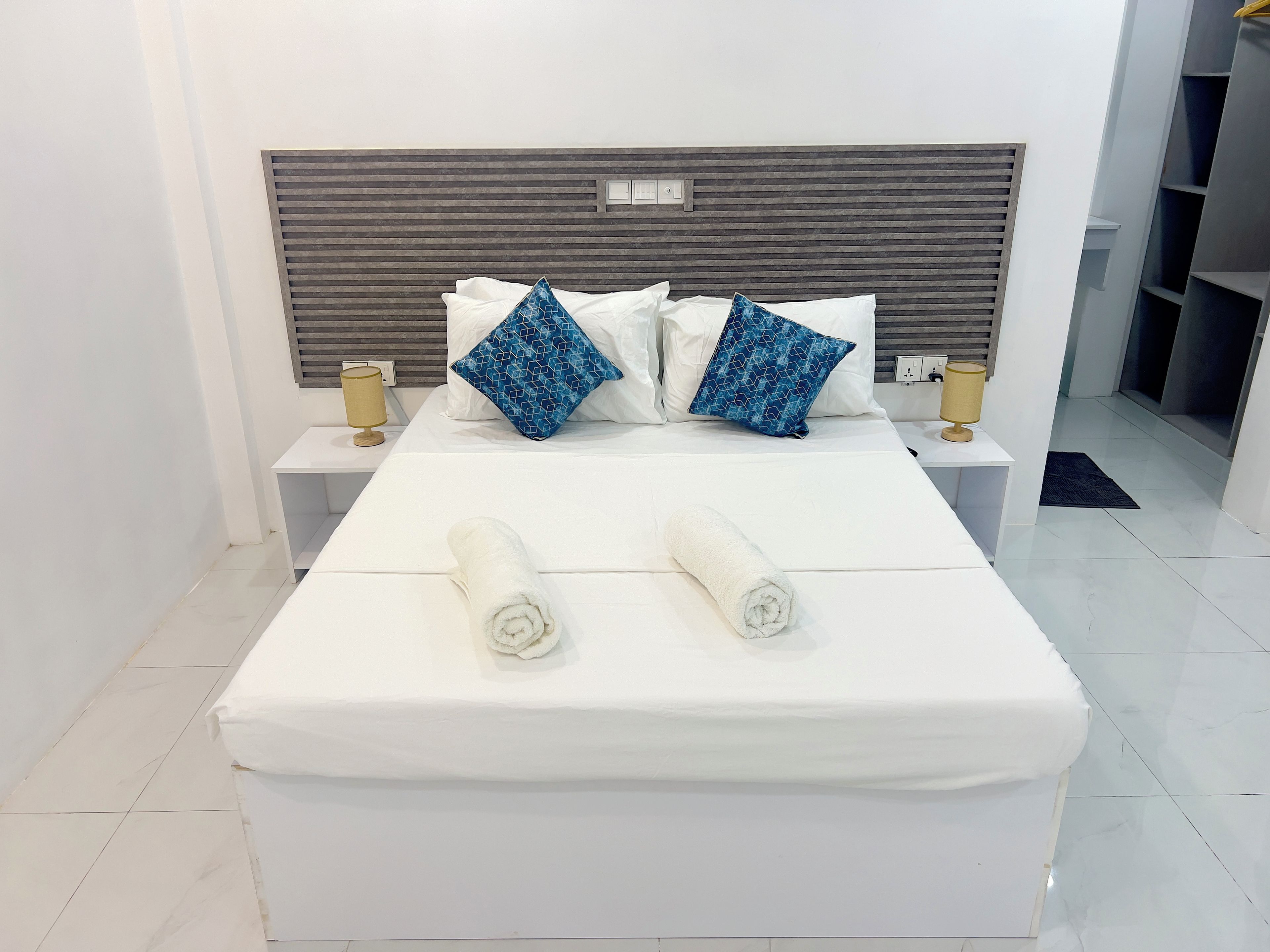 A neatly made bed with two pillows and two rolled towels, a bedside table with a lamp, and a headboard with a control panel.
