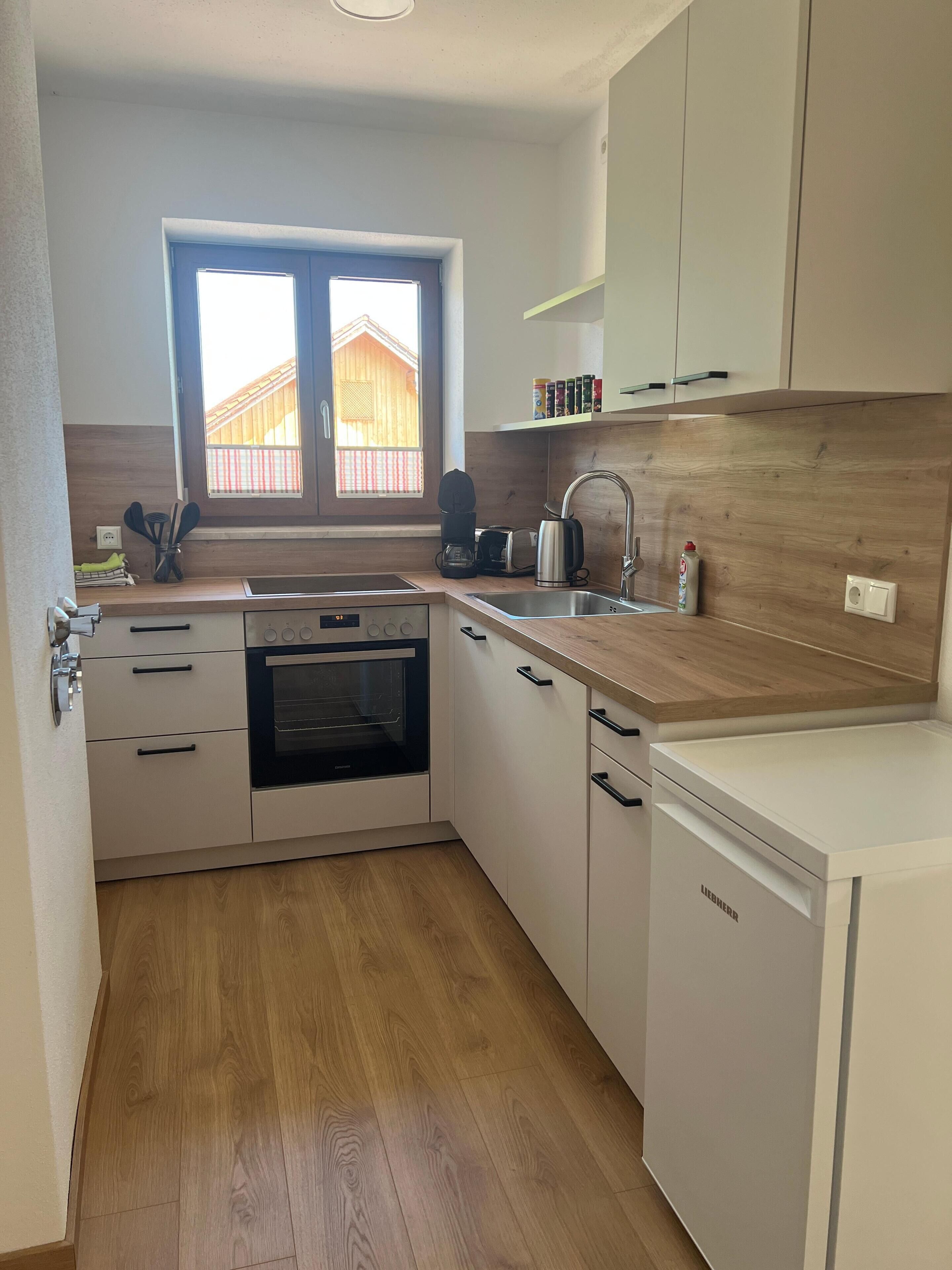 A compact kitchen with a built-in oven, a dishwasher, and a window with a view of a wooden structure outside.