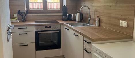 A compact kitchen with a built-in oven, a dishwasher, and a window with a view of a wooden structure outside.