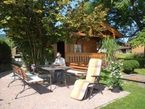 En utomhusplats med bord och stolar, en träkonstruktion med en veranda och en trädgård med en krukväxt.