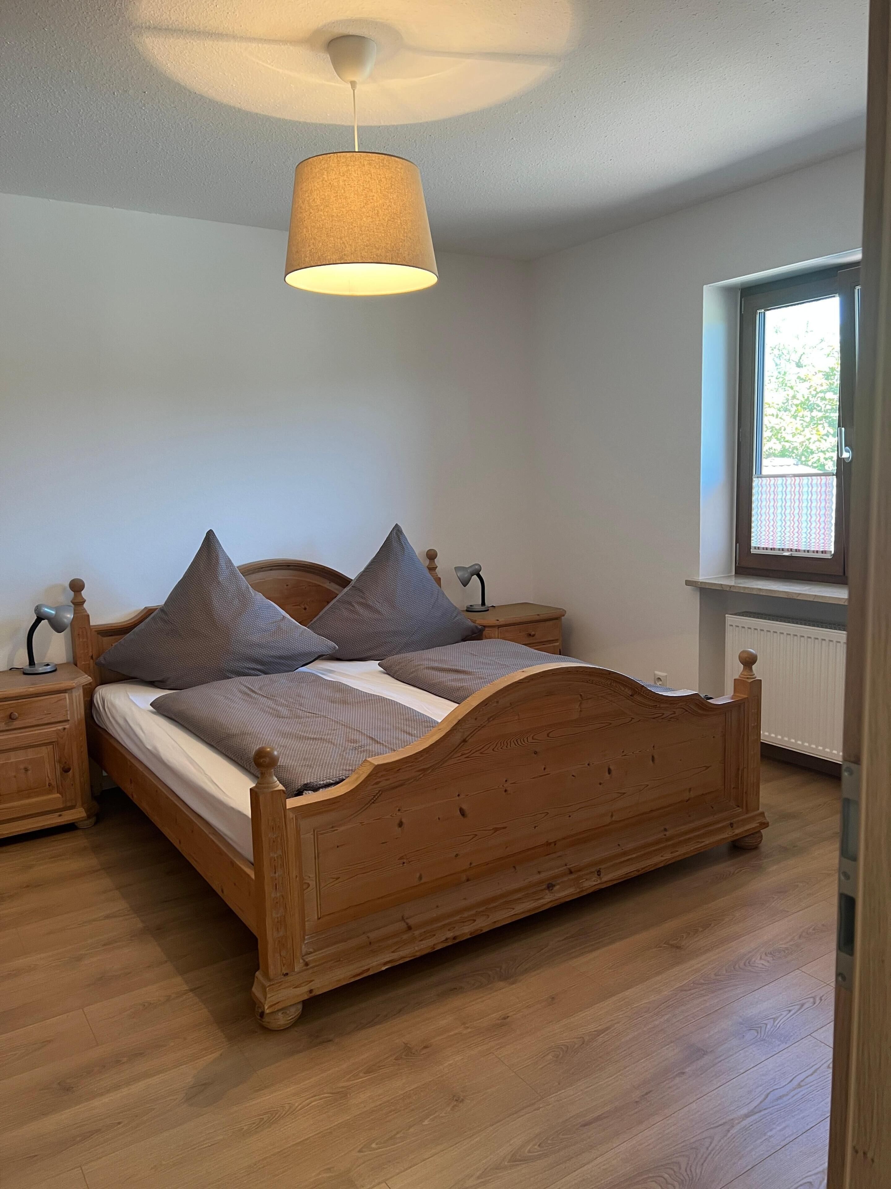 A bedroom with a wooden bed, two nightstands, a lamp, a window with blinds, and a ceiling light fixture.