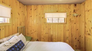 Une pièce aux murs lambrissés, avec un lit, une fenêtre équipée de stores et une porte dotée d’une serrure.