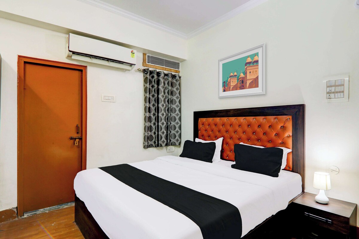 A hotel room with a bed, a nightstand, a lamp, an air conditioning unit, a door, and a wall-mounted painting.