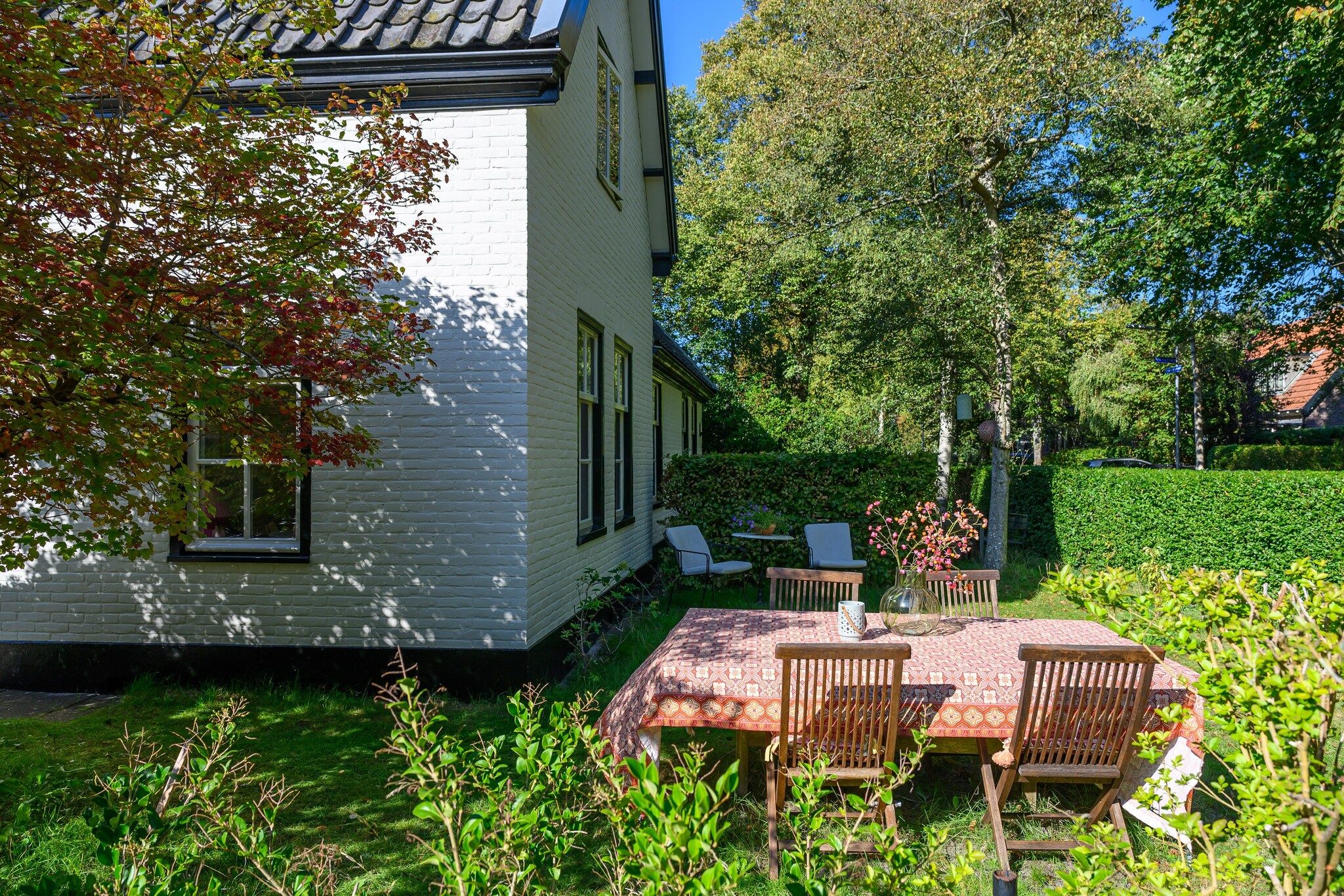 A white building with a brick facade, a patio with a patterned tablecloth and wooden chairs, and a well-maintained garden with various plants and a hedge.