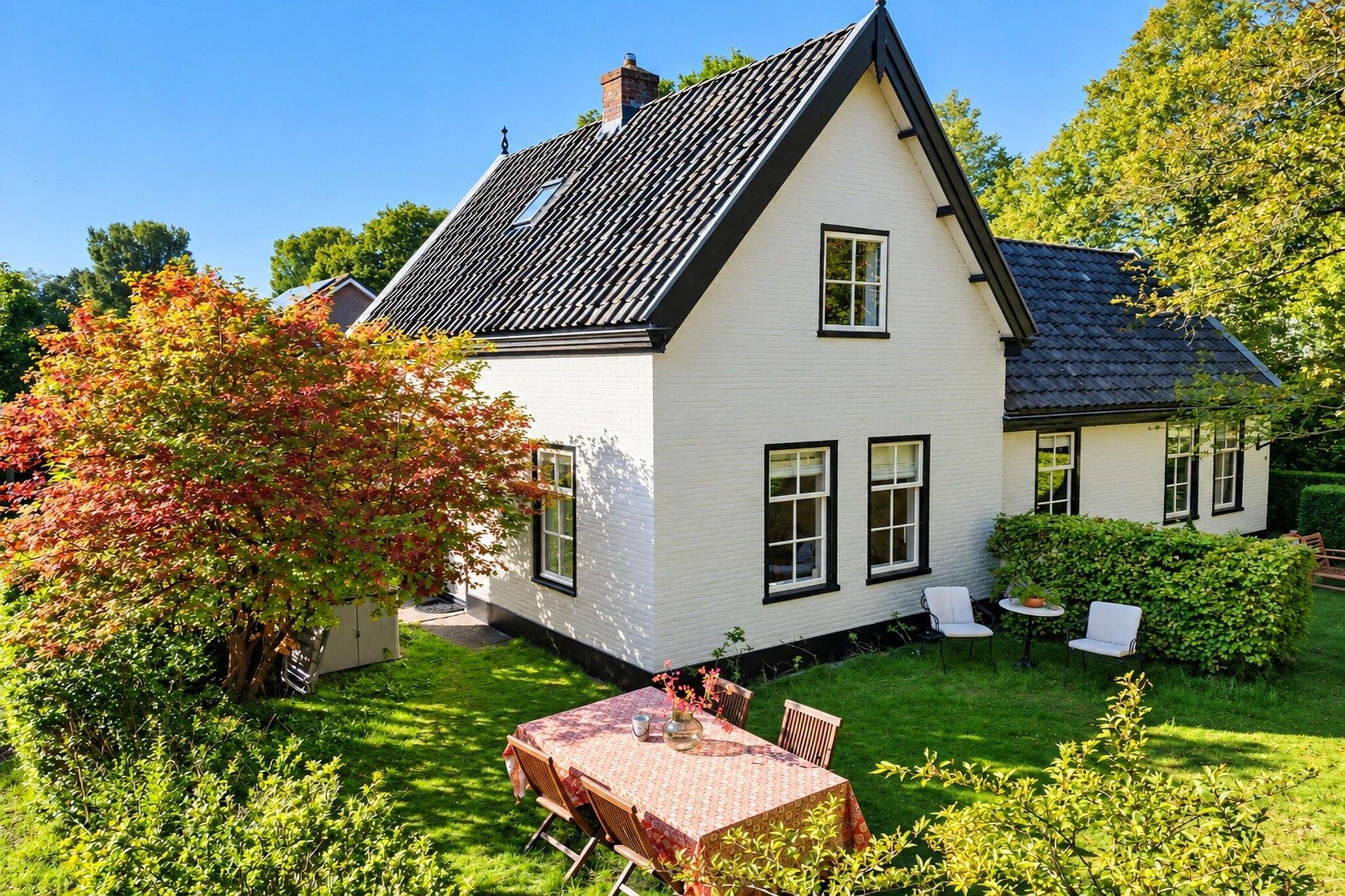 Et hvidt hus med sort tag, omgivet af en velplejet have og et spiseområde med bord og stole.