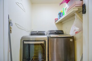 Pesuhuoneessa on kaksi päällekkäin asetettua pesukonetta ja kuivausrumpua, seinälle kiinnitetty hylly, jossa on siivoustarvikkeita, sekä seinällä roikkuva muovipussi.