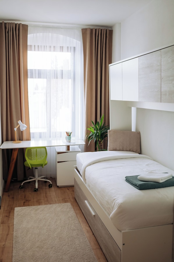 A bedroom with a bed, desk, chair, and a window with curtains.