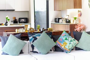 A modern living room with a sofa adorned with patterned cushions, a dining area with a wooden table, and a kitchen in the background. - Villa Koa 4BR at Aileen Villas Layan (Choeng Thale)