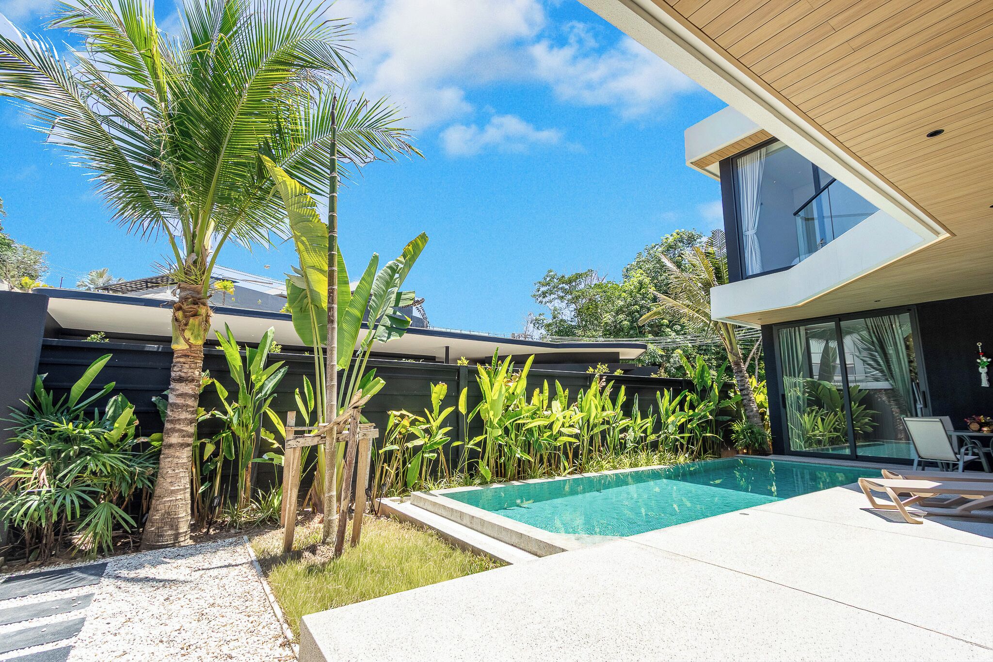 Un área de piscina moderna al aire libre con un cielo azul claro, rodeada de plantas tropicales y un edificio contemporáneo.
