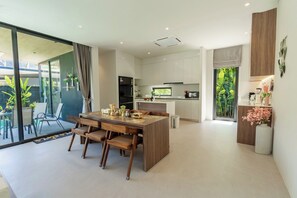 A modern kitchen with a dining area, large windows, and a view of greenery. - Villa Koa 4BR at Aileen Villas Layan (Choeng Thale)