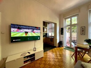 A living room with a flat-screen TV mounted on the wall, a wooden floor, a dining table with chairs, and a sliding glass door leading to an outdoor area. - New Villa Apartment, Sleeps 6, Balcony, Ideal for Football Teams Schalke 04! (Gelsenkirchen)