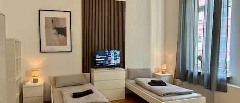 A modern hotel room with a bed, bedside tables, a TV, and a window with curtains.