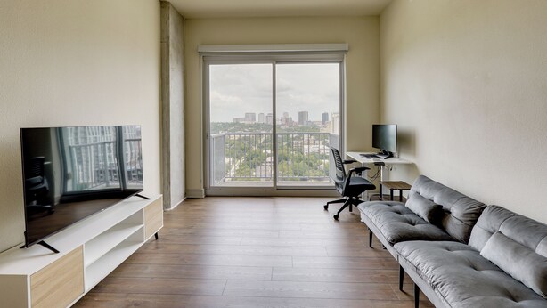 Una sala de estar moderna con una televisión de pantalla plana, un sofá gris, un escritorio con una computadora y un ventanal con vistas a la ciudad.