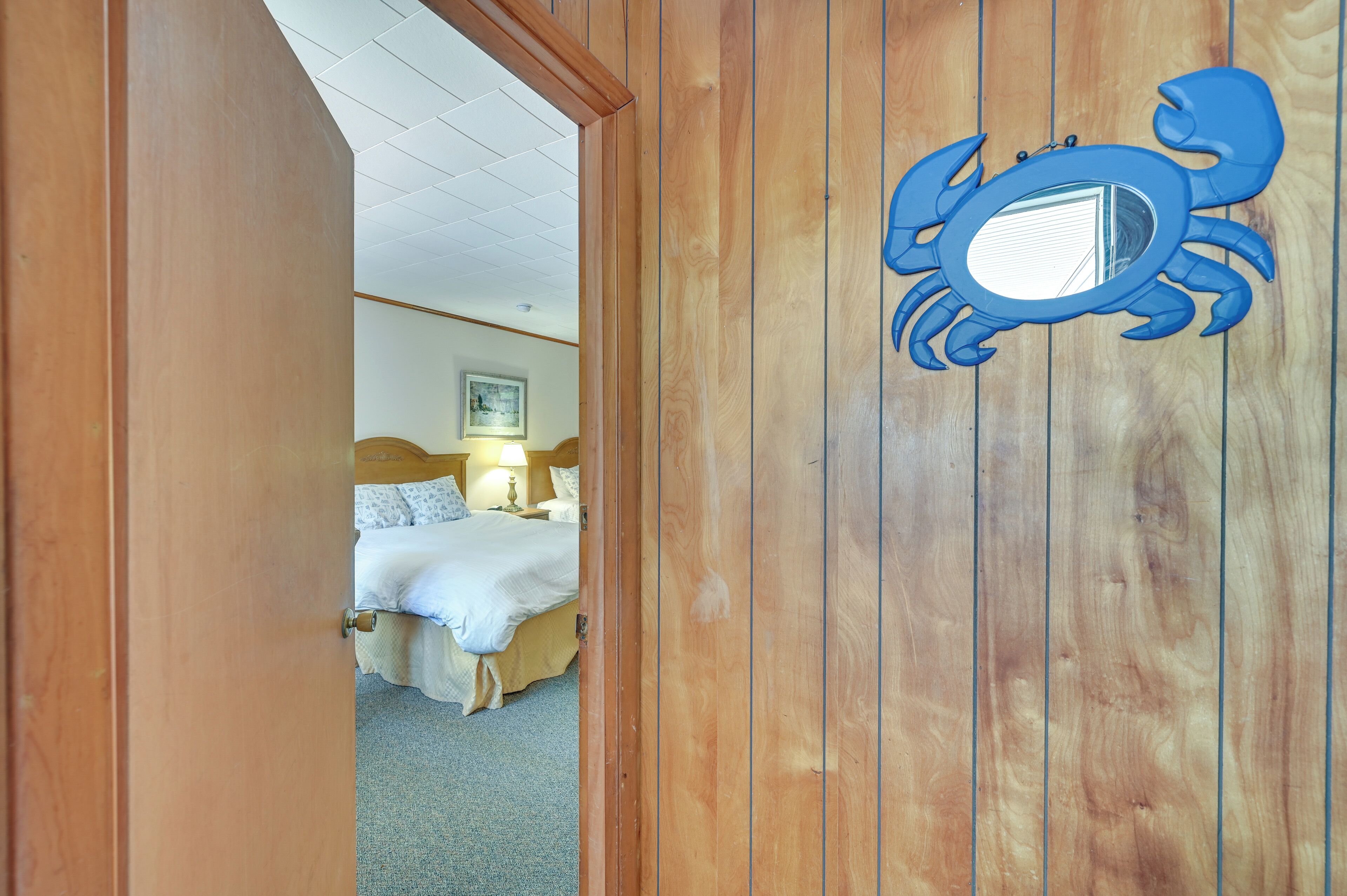 A wooden door with a blue crab decoration, leading to a bedroom with a bed and a nightstand.