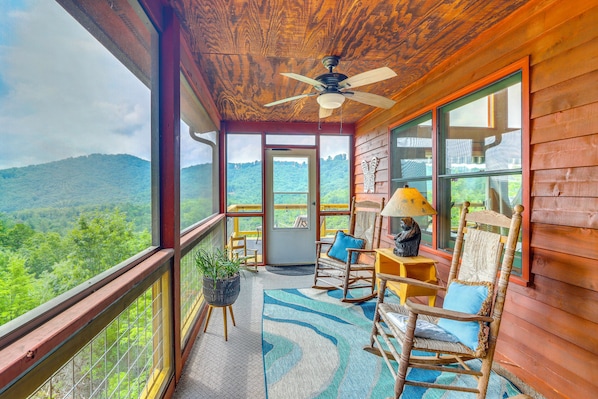 A wooden balcony with a ceiling fan, a view of a forested landscape, and two rocking chairs with cushions. - Stunning Views & Sunrises! Pappy's Place in Murphy (Murphy)
