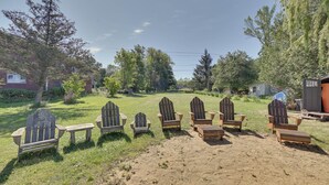 Een rij Adirondack-stoelen en een bank in een groene buitenruimte, met bomen en een huis op de achtergrond.